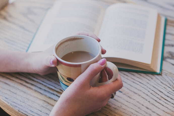 Pessoa segurando caneca na mesa junto de um livro