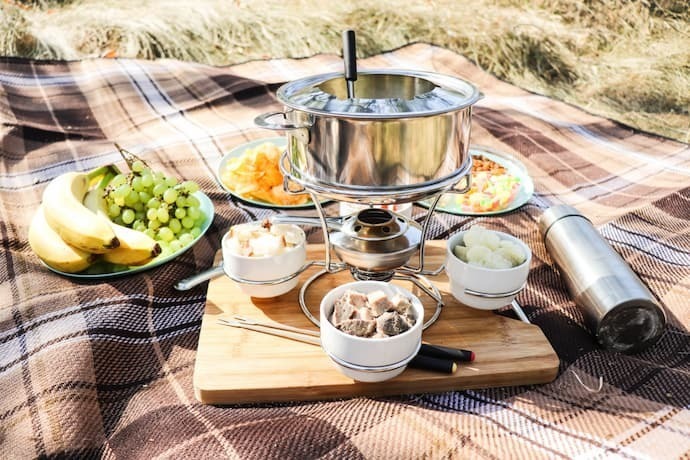 Piquenique com fondue de alumínio e com frutas ao redor.