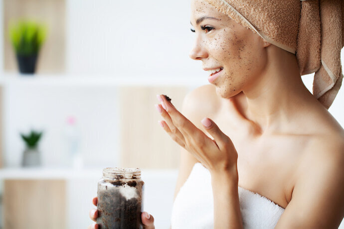 Mulher aplicando esfoliante no rosto no banheiro e segurando o pote do produto.