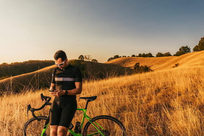 Homem encostado em sua bicicleta, mexendo no celular e atrás a vista de um campo