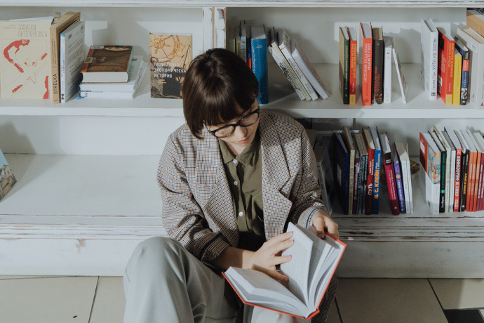 Indivíduo lendo um livro e ao fundo uma estante de livros enfileirados e organizados