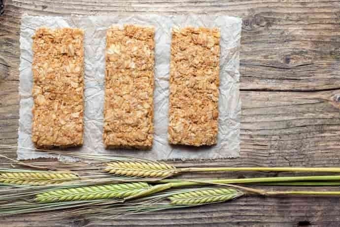 Apresenta-se na imagem um papel, e sobre ele temos três barras de cereais afastadas uma da outra. Além disso, abaixo das barras de cereais estão dispostas quatro gramíneas de trigo. 